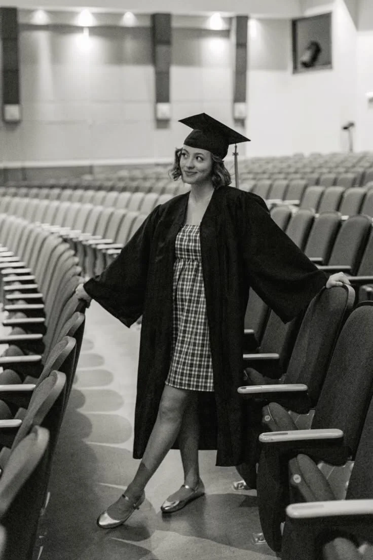 A woman in a graduation cap and gown standing in an auditorium with empty seats, smiling and posing with arms outstretched.
