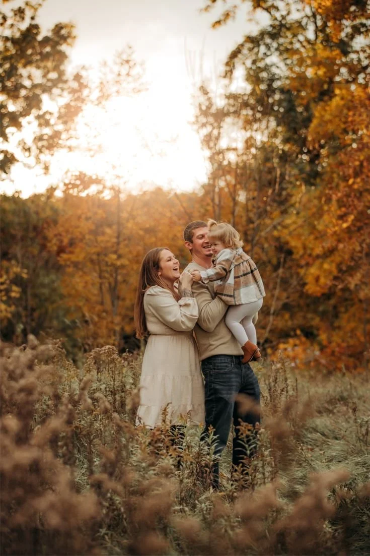 A happy family of three enjoying fall outdoors, with orange and yellow trees in the background and warm sunlight.