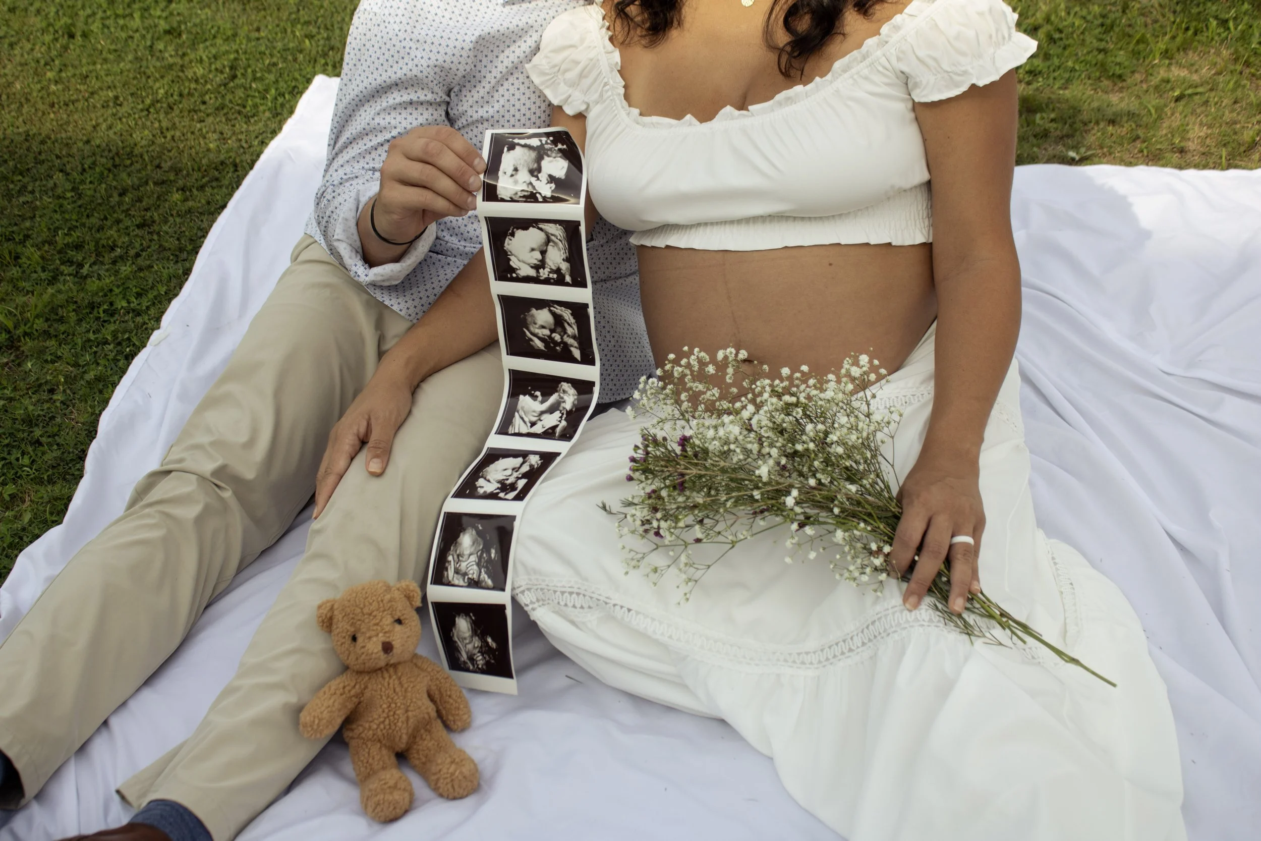 A pregnant woman lying on her side on a white blanket outdoors, holding a 3D ultrasound image of her baby. She has a flower bouquet on her belly and a teddy bear resting next to her. Her partner is sitting behind her, holding the ultrasound image.