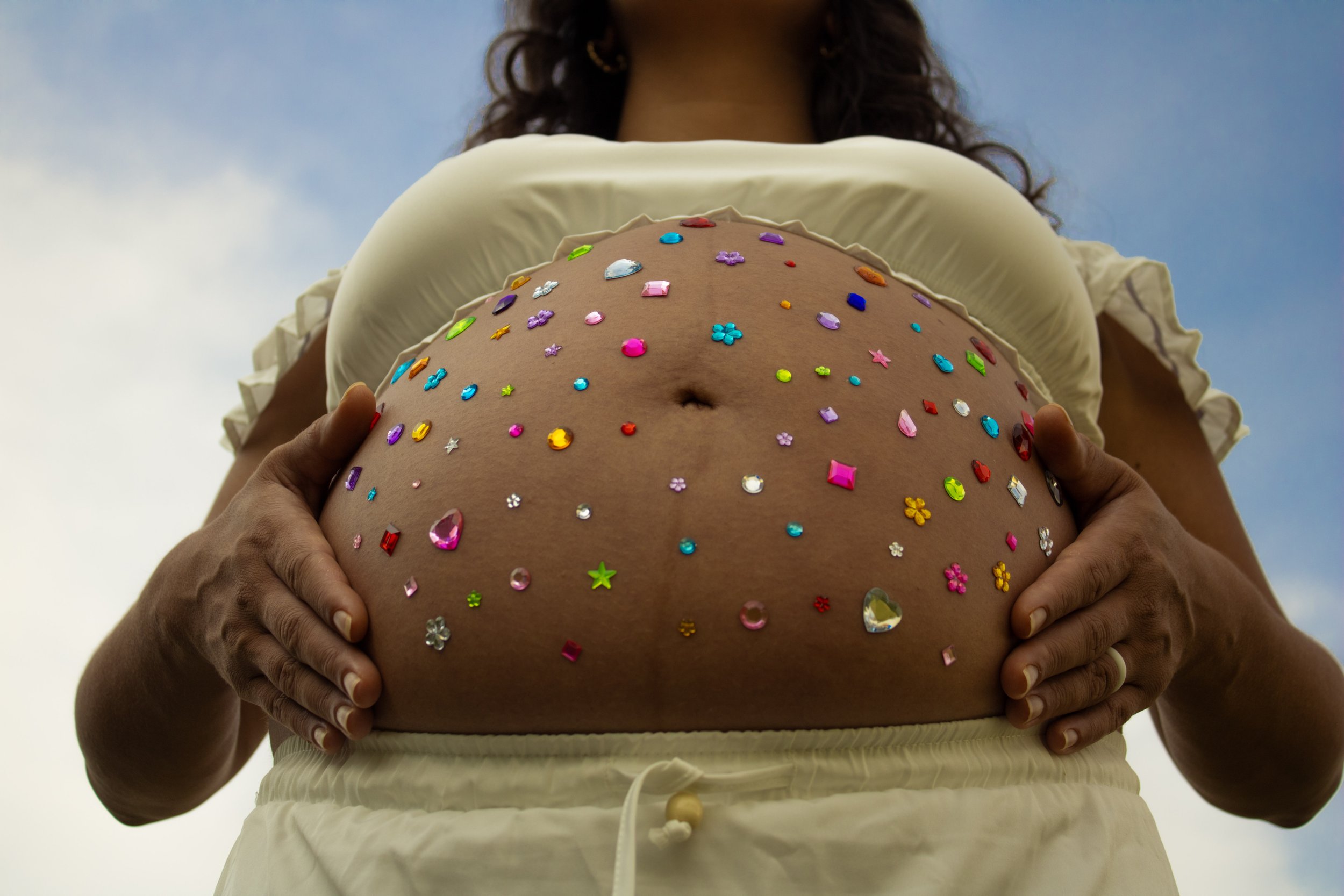 Back view of a woman wearing a cream-colored top, holding her pregnant belly, which is decorated with colorful gemstone and sticker embellishments against a cloudy sky background.
