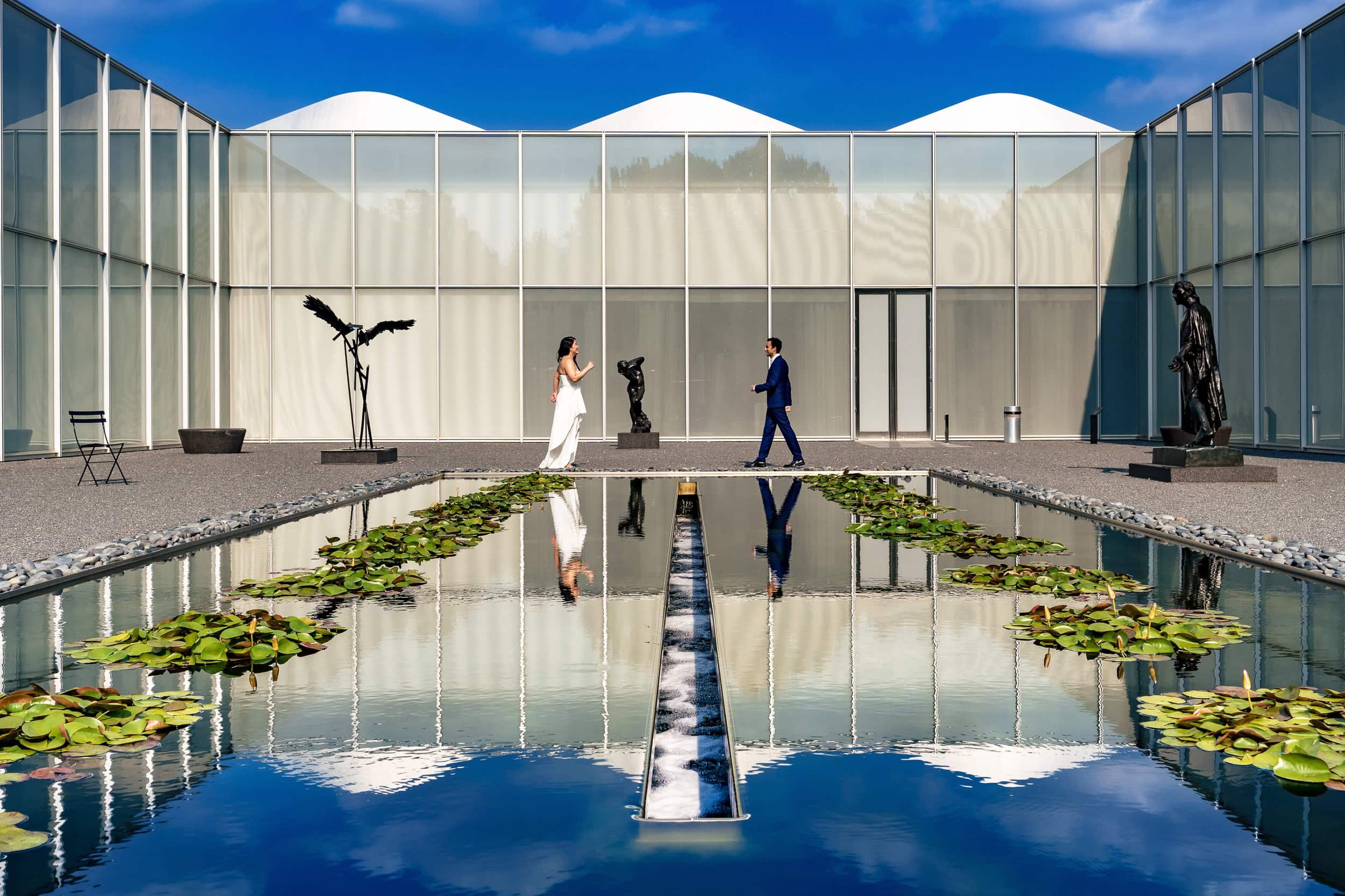 A modern building with glass walls and a white roof, outdoor courtyard with a rectangular pond filled with lily pads, a woman and man are talking near sculptures, and the sky is partly cloudy.