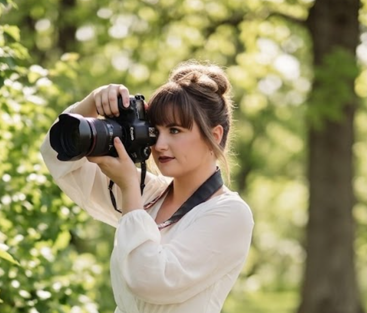 Young woman with brown hair and bangs, wearing a white blouse, holding a camera up to her eye, standing outdoors in a green, wooded area.