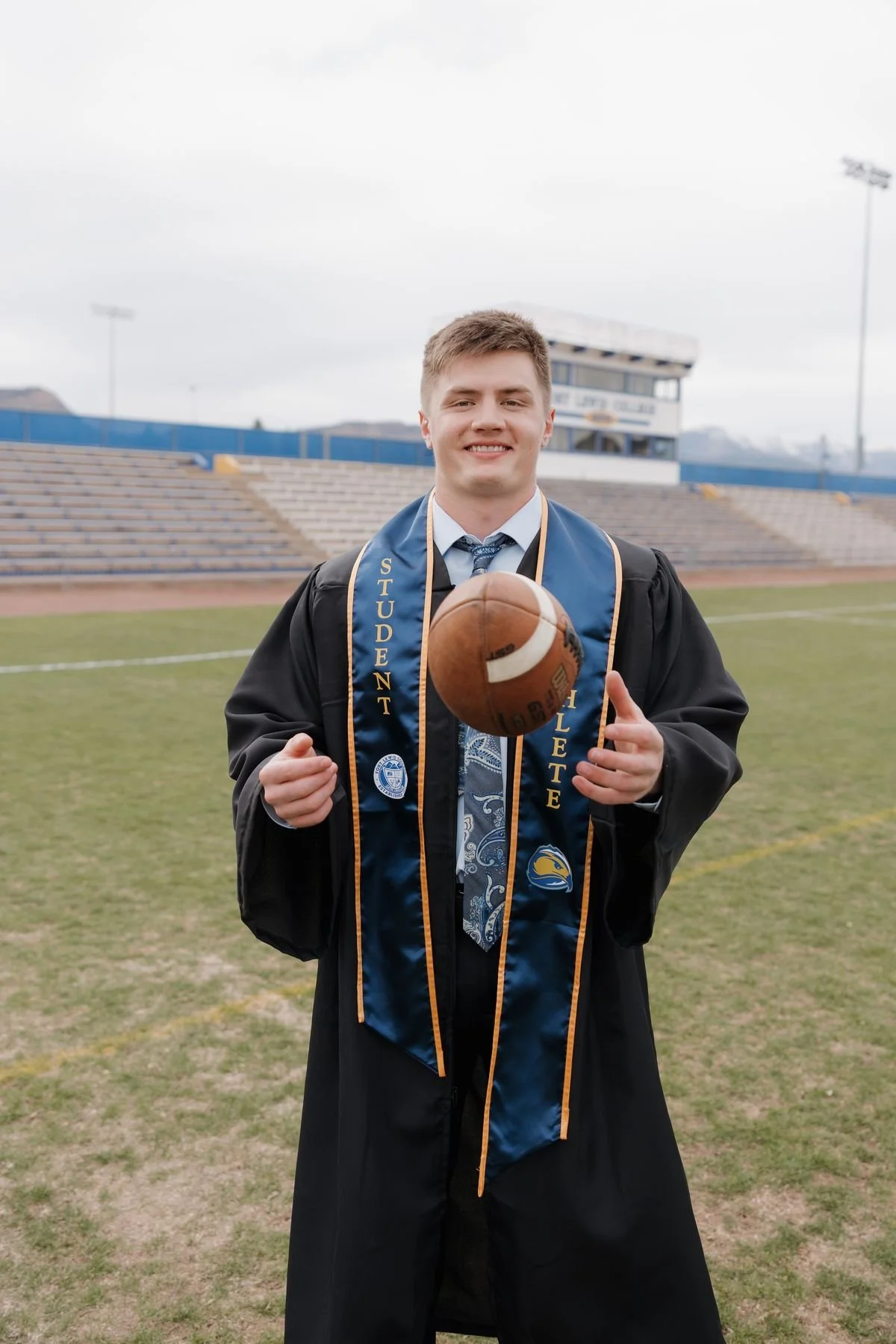 A young man in a graduation gown and cap standing on a football field, smiling, holding a football in front of him with both hands. He wears a blue stole that reads 'STUDENT ATHLETE' and has a school logo, with bleachers and the stadium structure in the background.