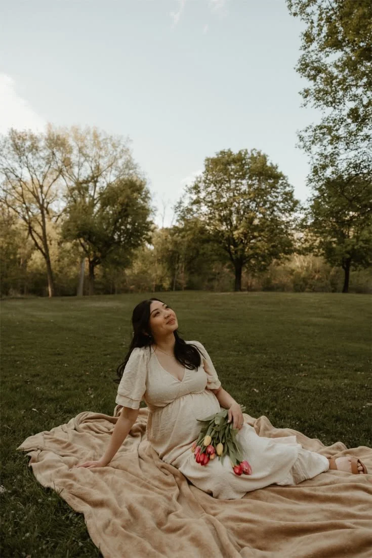 A woman in a cream-colored dress sitting on a blanket in a park, holding a bouquet of pink tulips, with trees and blue sky in the background.