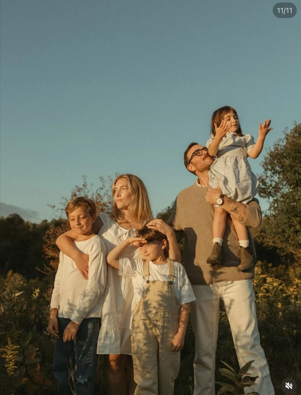 A family of six standing outdoors at sunset, with children and adults, some looking into the distance and others interacting, surrounded by trees and plants.