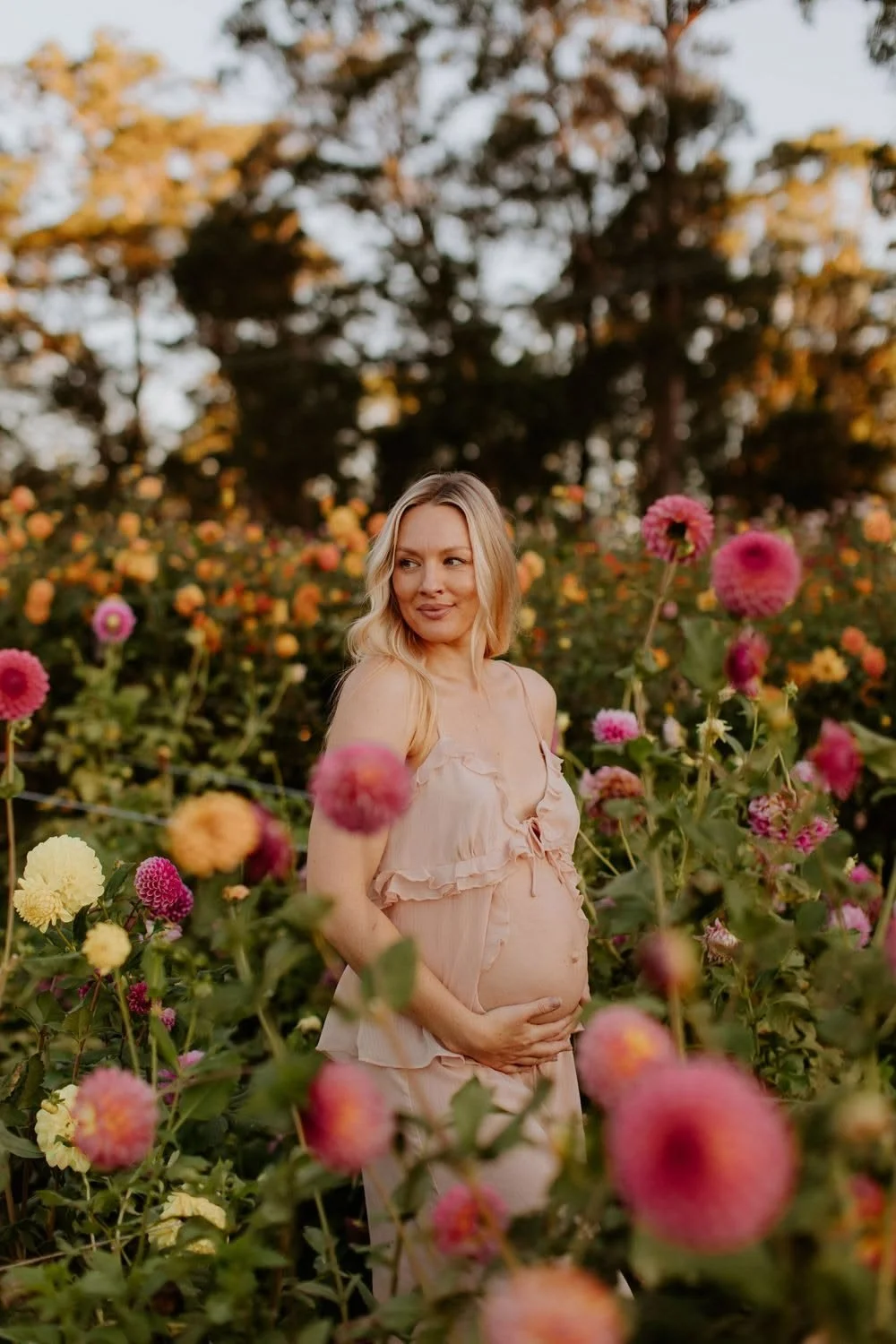 A pregnant woman standing in a colorful flower garden, gently holding her baby bump, with trees in the background during sunset.