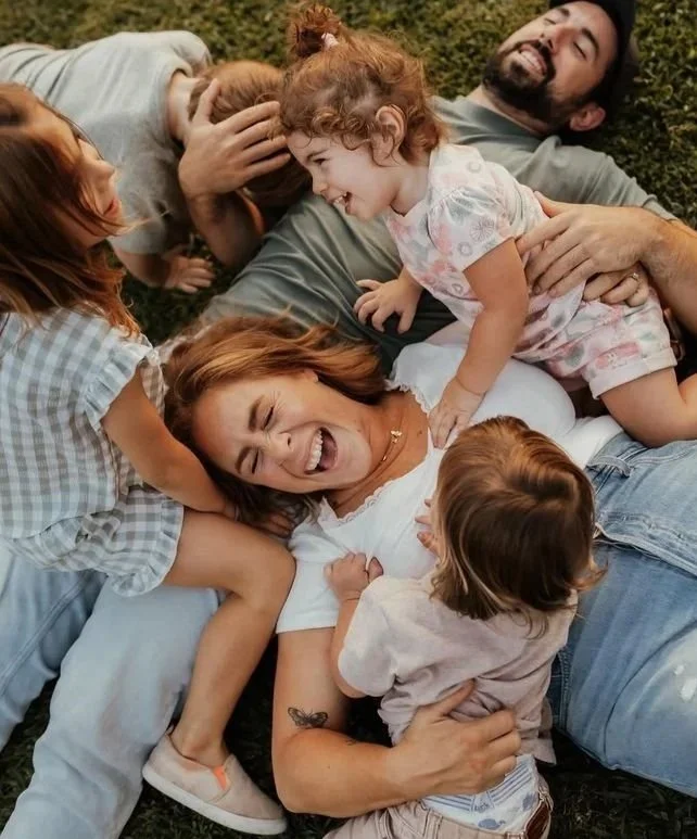 A joyful family of six, including three adults and three children, is playfully lying on the grass, smiling and laughing together.