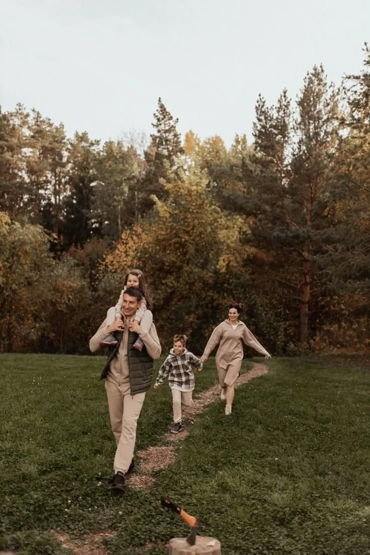 Family of four walking and playing outside on a trail in a forested area during autumn, with trees with green and orange leaves in the background.