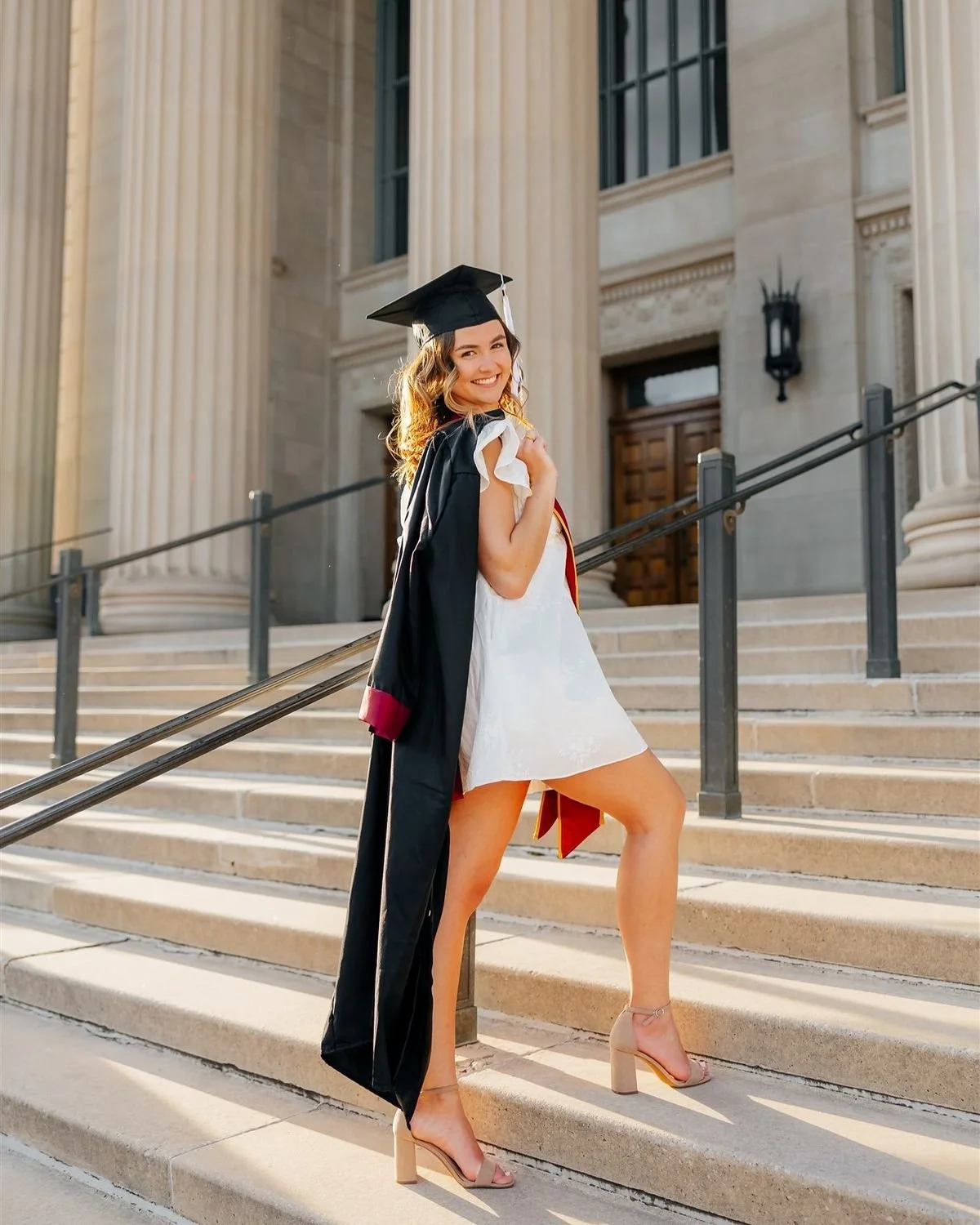 A young woman in a white dress and high heels is celebrating her graduation on the steps of a large, classical building, holding a graduation cap and gown.