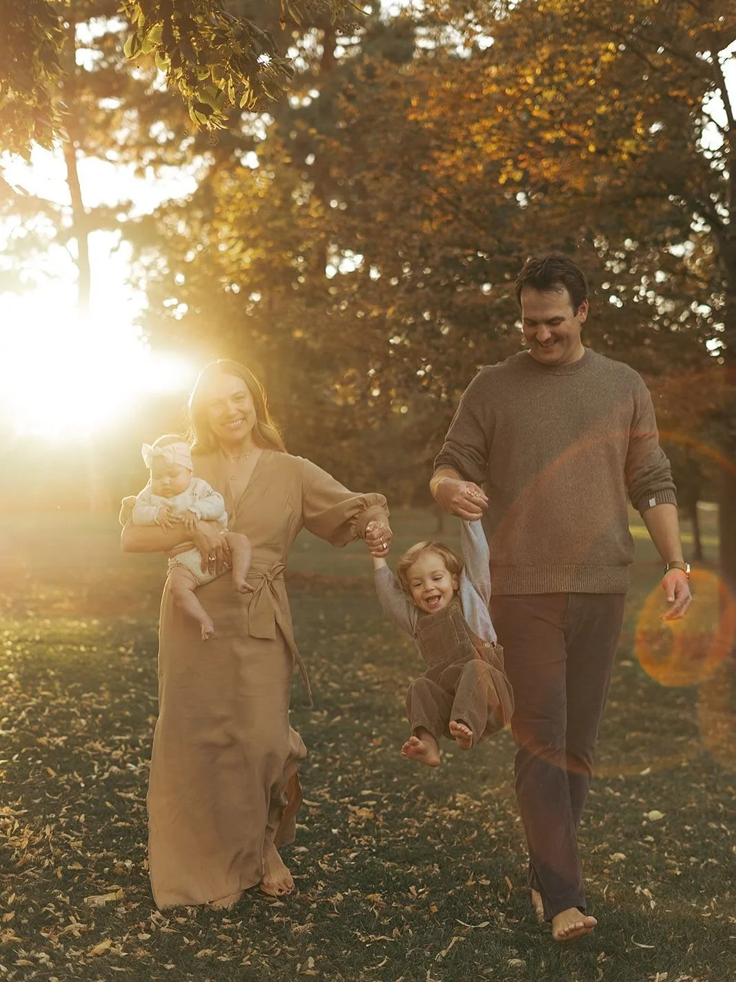 A family of four enjoying a walk in a park during autumn sunset, with the mother carrying a baby and the father holding a young child's hand as they play and smile.