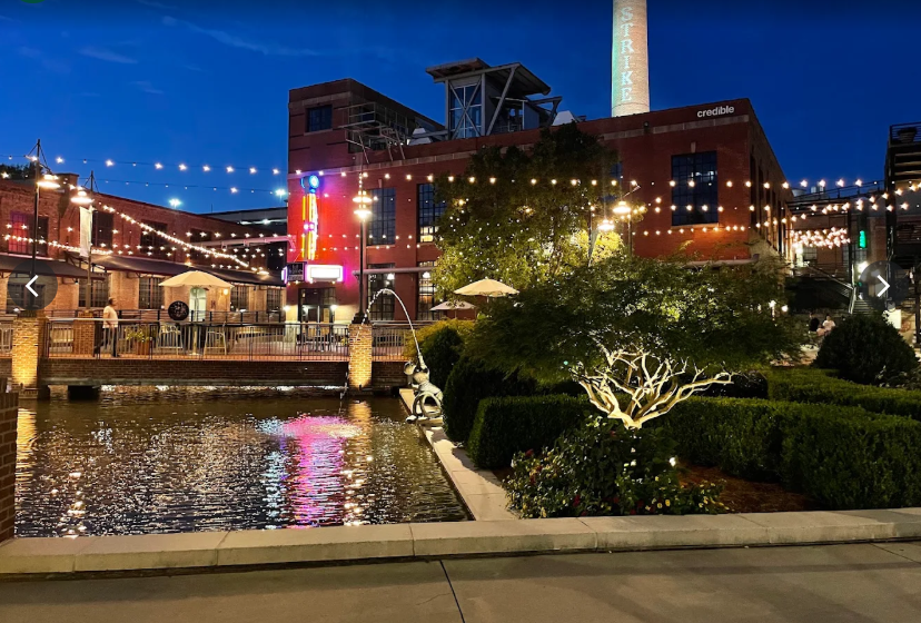 Night view of an outdoor area with string lights, a water feature, and a brick building with illuminated signage and smokestack in the background.