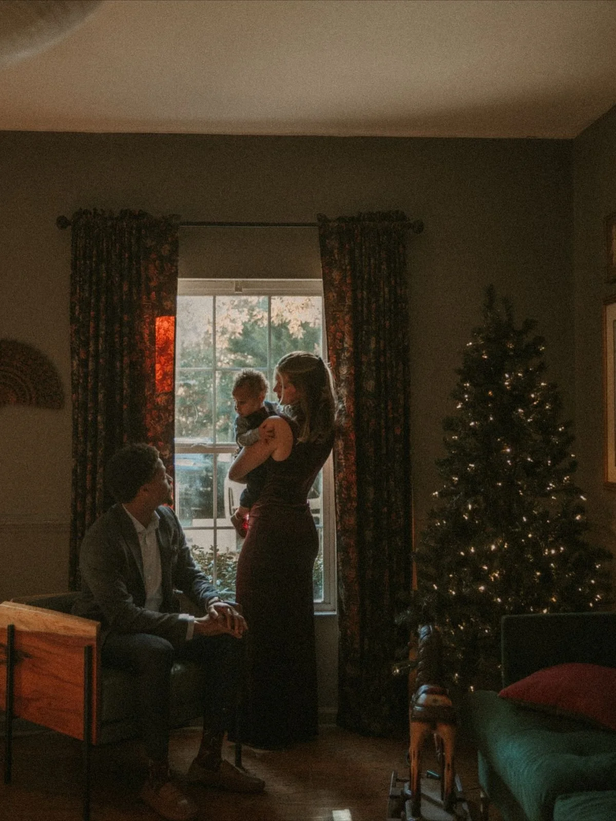 A family gathers near a window with curtains, with a decorated Christmas tree to the right, during a festive evening.