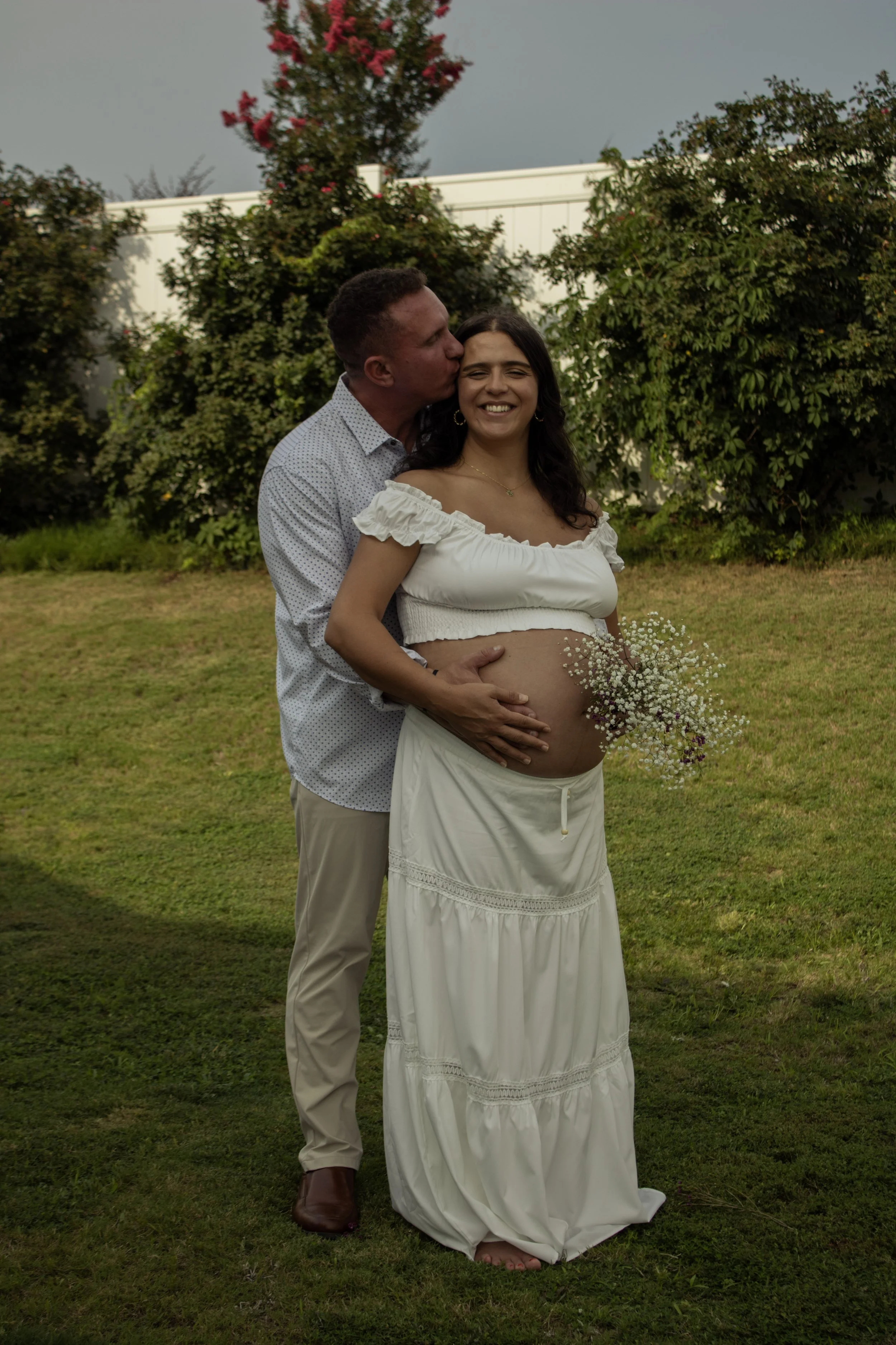 A couple standing outdoors on grass, with a woman revealing a pregnant belly, holding a bouquet of small white flowers, and a man kissing her on the cheek, both smiling, background of trees and a white fence, overcast sky.