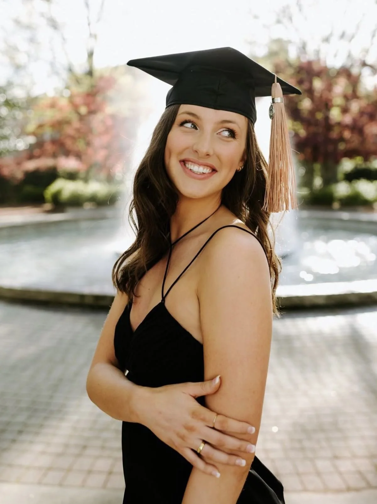 Young woman in a black dress and graduation cap stands outdoors near a fountain, smiling and looking to her right.