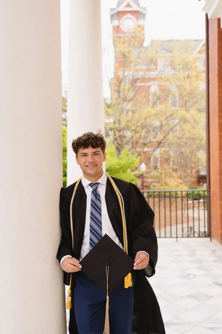 Young man in a graduation gown and cap standing outdoors next to a white pillar with a historic building with a clock tower in the background.