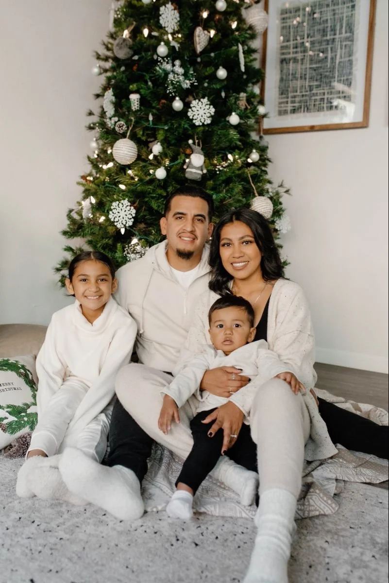 A family of four sitting on the floor in front of a decorated Christmas tree, with a man, woman, and two children smiling indoors.