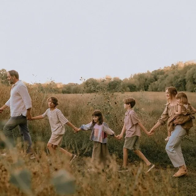 A family walking hand in hand through a grassy field with trees in the background during sunset.