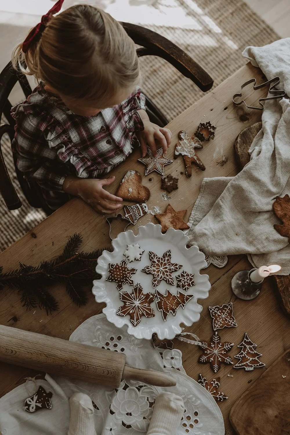 A young girl in a plaid shirt sits at a wooden table decorating Christmas cookies, including star, snowflake, and Christmas tree shaped cookies, with icing and sprinkles.