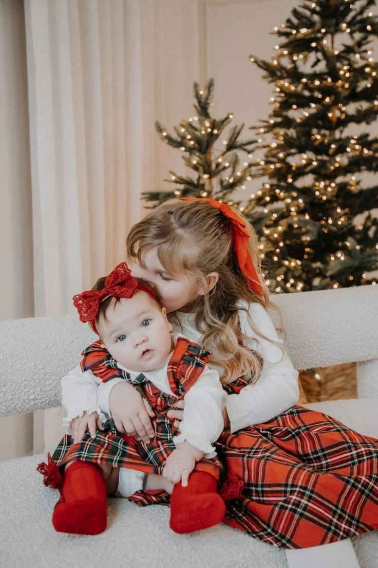 A young girl hugging a baby girl, both dressed in red and tartan Christmas outfits, sitting on a white bench with decorated Christmas trees in the background.