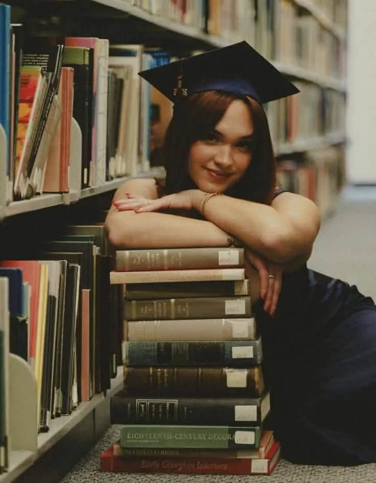 A woman with brown hair wearing a graduation cap is leaning on a large stack of books in a library.