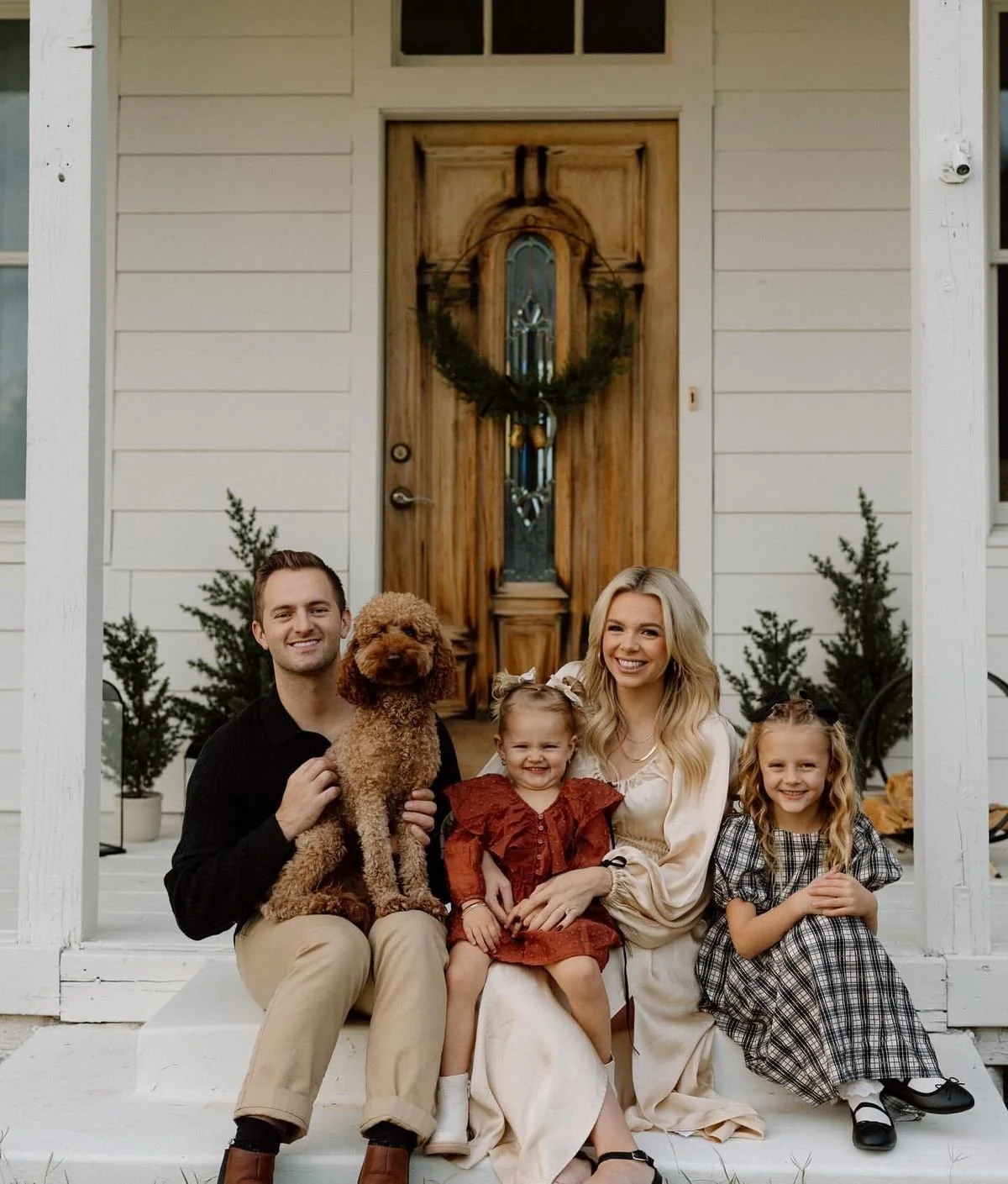Family sitting on front porch of house with wooden door and wreath, mom, dad, two young girls, and a dog.