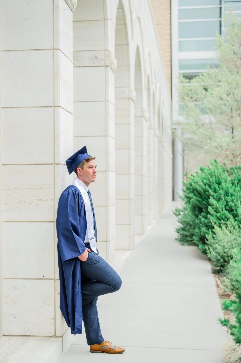 A young man in a blue graduation gown and cap leaning against a stone building on a sidewalk, with greenery and modern buildings in the background.