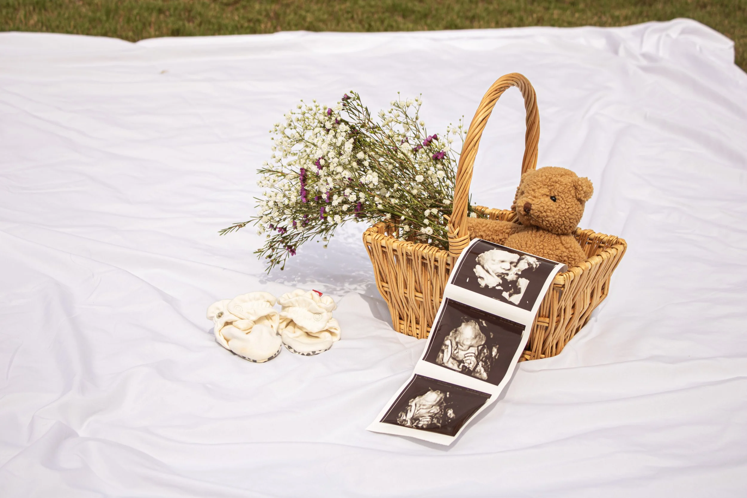 A wicker basket with a teddy bear, surrounded by white and purple flowers, ultrasound images, and baby socks on white fabric outdoors.