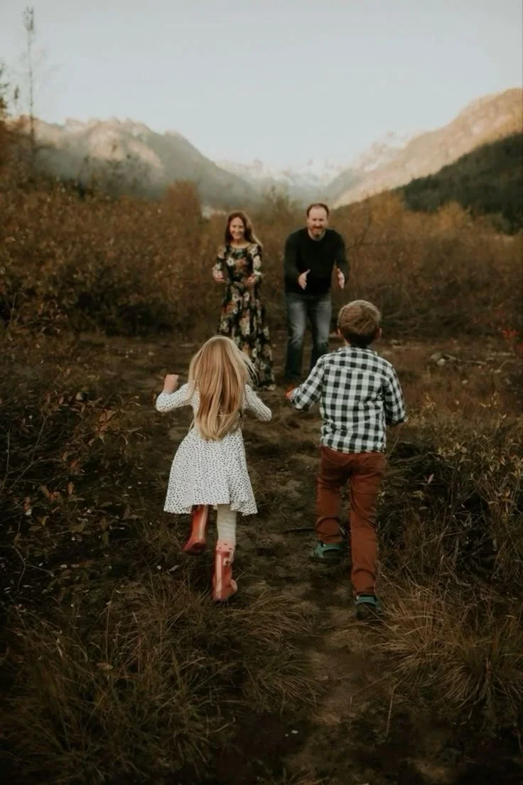 A family of four running along a dirt trail in a mountainous, autumn landscape. The children, a girl in a polka dot dress and rain boots and a boy in a plaid shirt and brown pants, lead the way, while the parents follow behind smiling in the distance.