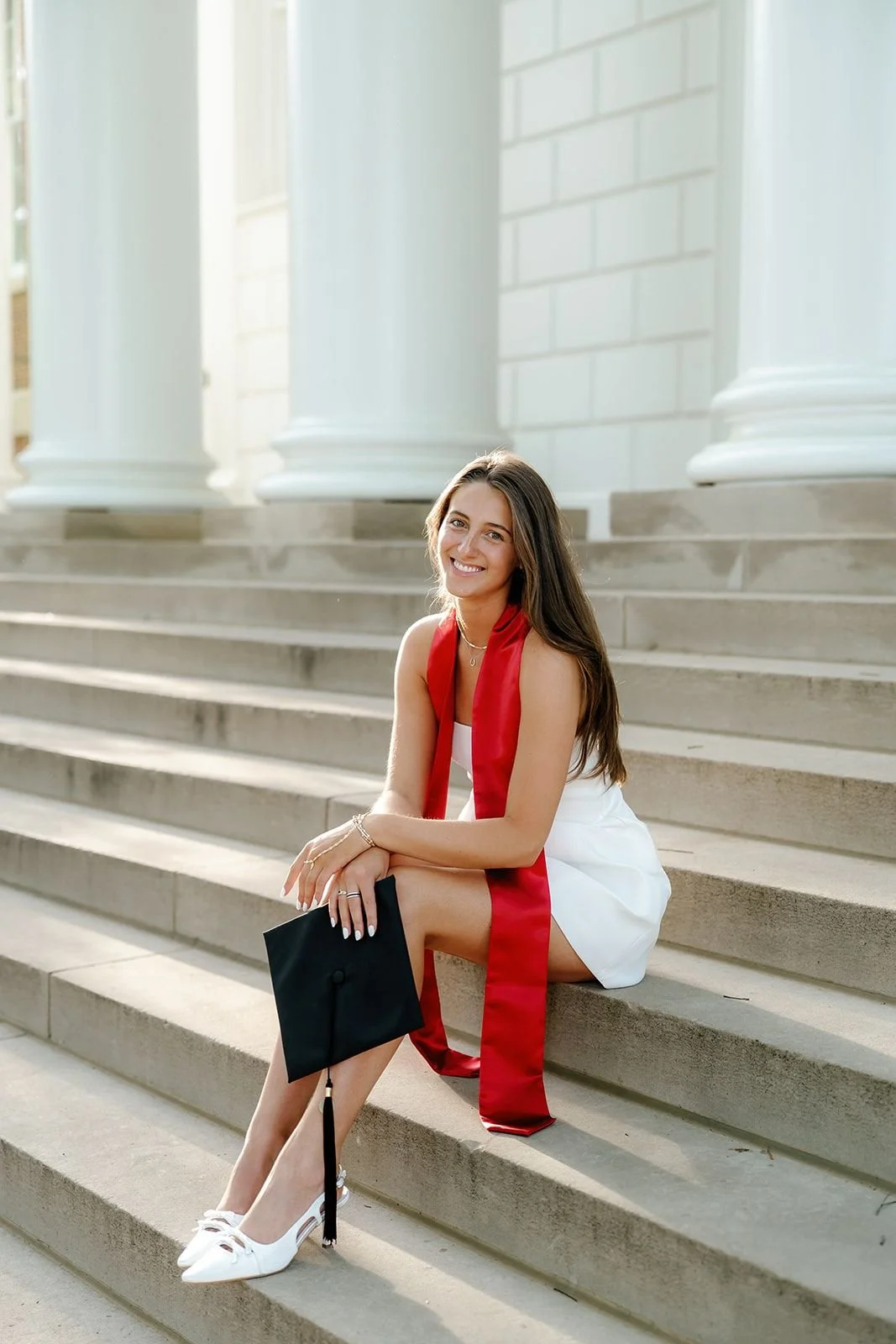 A young woman in a white dress with a red sash, holding a black graduation cap, sitting on the steps in front of a white building with large columns, smiling.