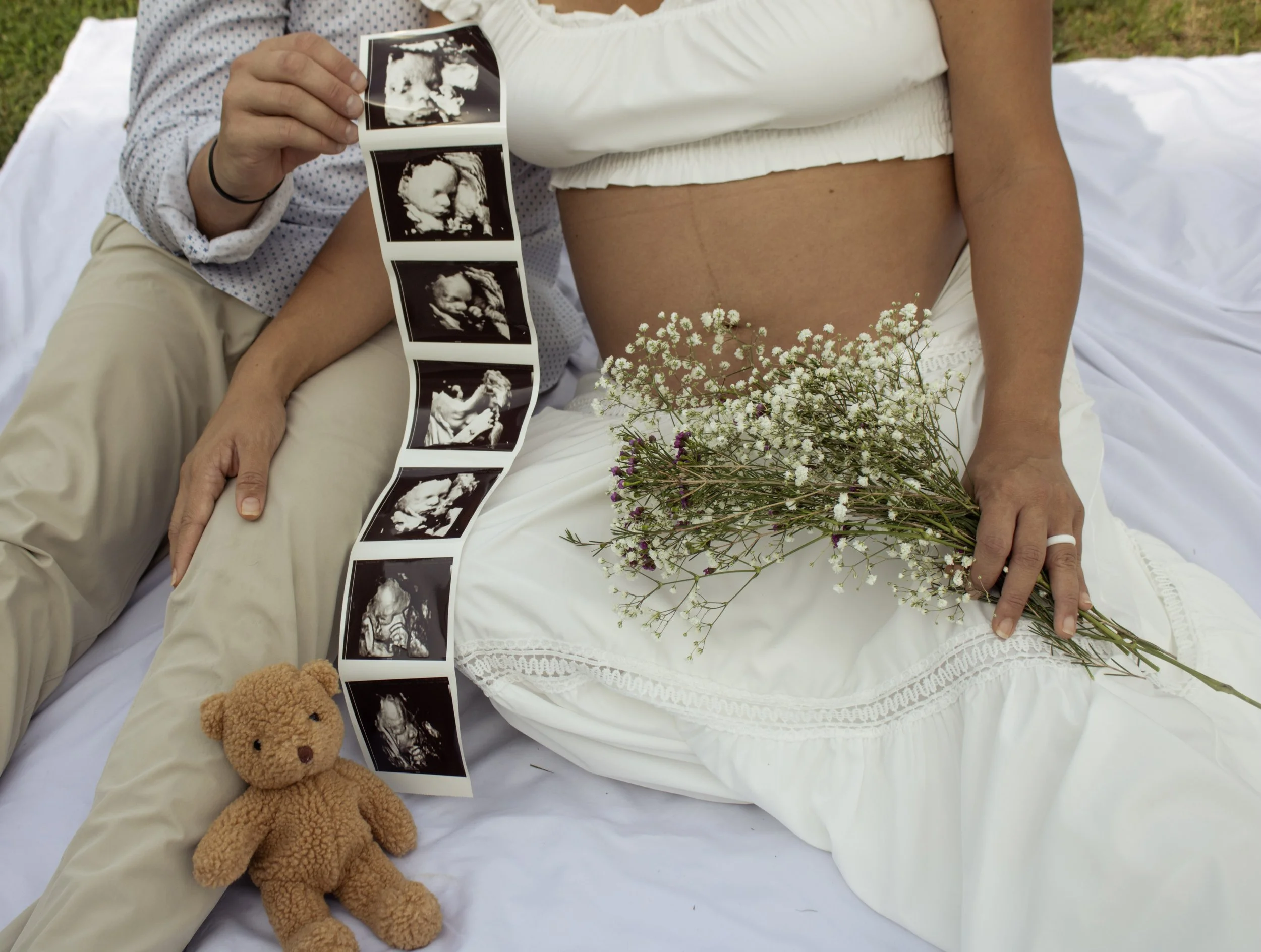 A pregnant woman lying on a bed holding a series of ultrasound images, with a bouquet of small white flowers resting on her belly, and a teddy bear placed on the bed near her.