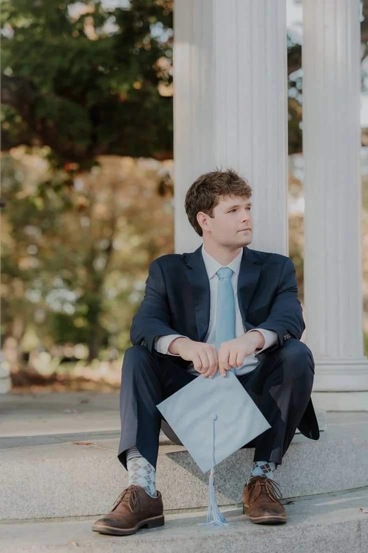 Young man in a dark suit sitting on steps outside, holding a graduation cap, with white columns in the background and trees with fall leaves.