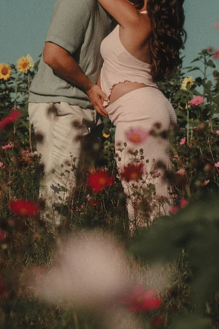 A couple standing close together in a field of colorful flowers, with the man gently holding the woman’s waist and the woman wearing a light-colored dress with curly hair.