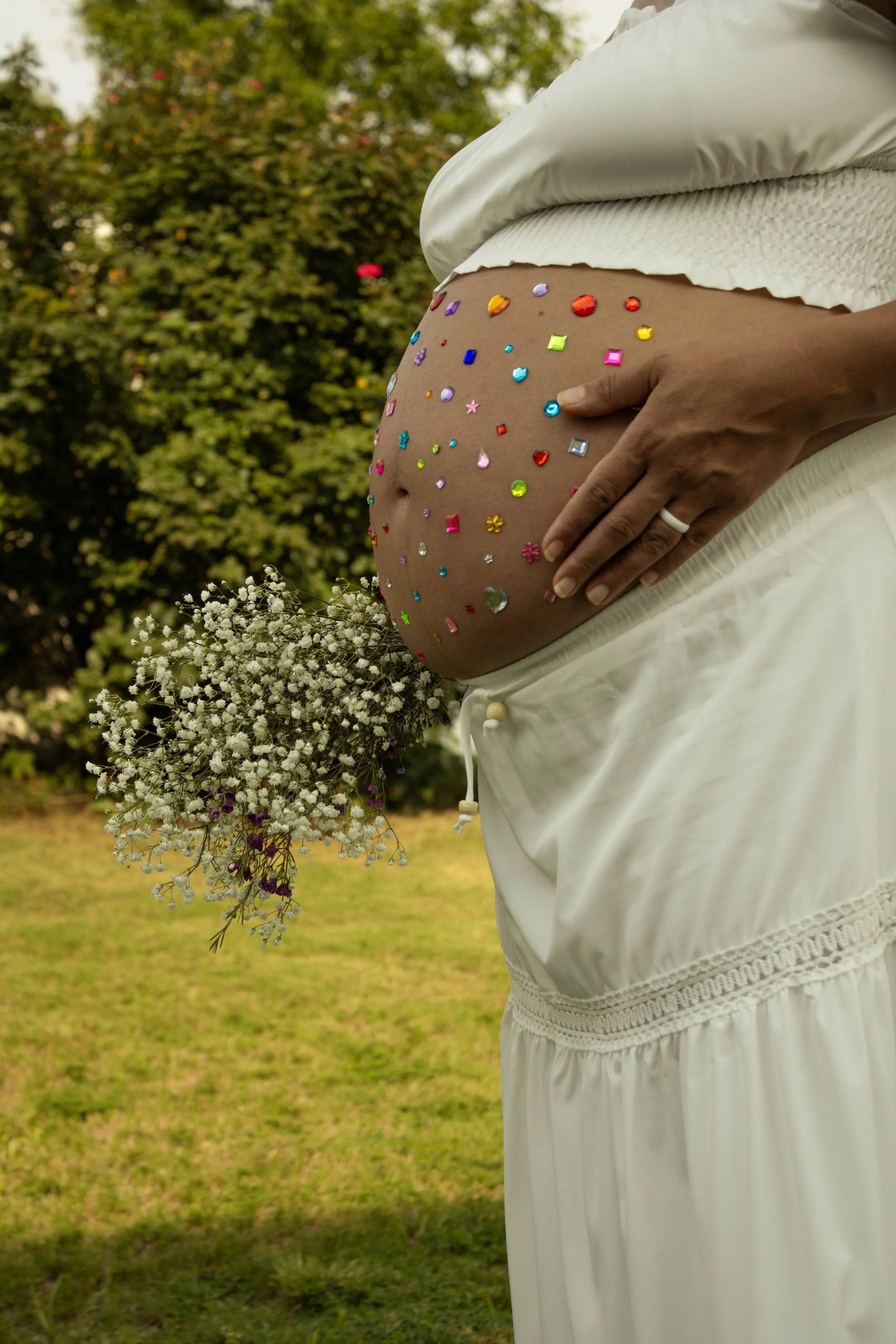 Close-up of a pregnant woman in a white dress with colorful rhinestones on her belly, holding a small bouquet of white baby's breath flowers, in an outdoor setting with green trees and grass in the background.