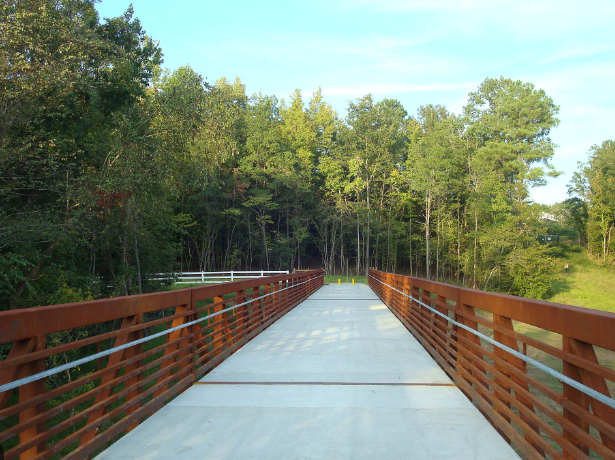 Empty concrete bridge with brown wooden railings, surrounded by trees and greenery under a blue sky.