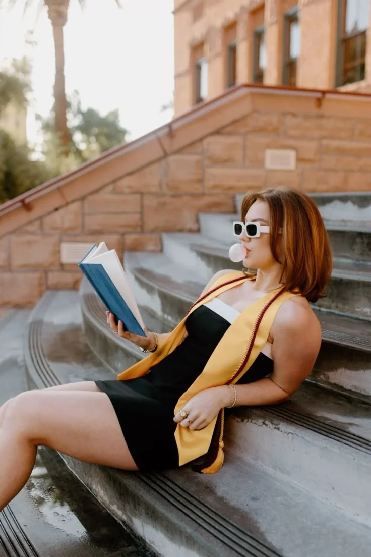 A woman with red hair, wearing sunglasses and a black dress with a yellow vest, sitting on outdoor concrete stairs, blowing a bubble with gum, reading a book.