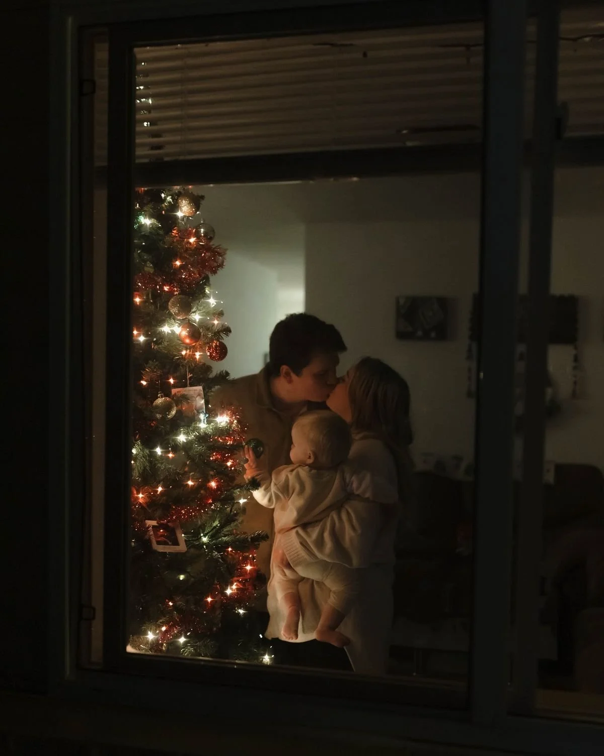 Three people, two adults and a young child, sharing a kiss in front of a decorated Christmas tree with lights and ornaments.
