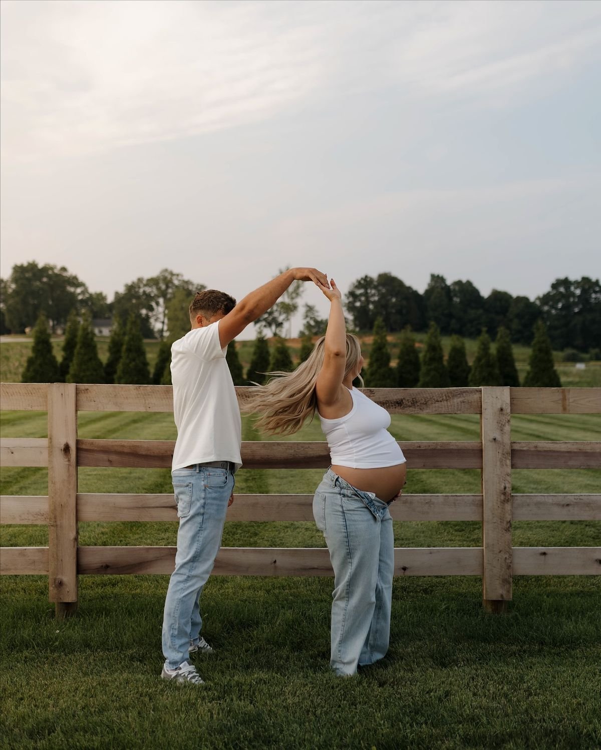 A pregnant woman and a man are dancing outdoors behind a wooden fence, with trees and a cloudy sky in the background.