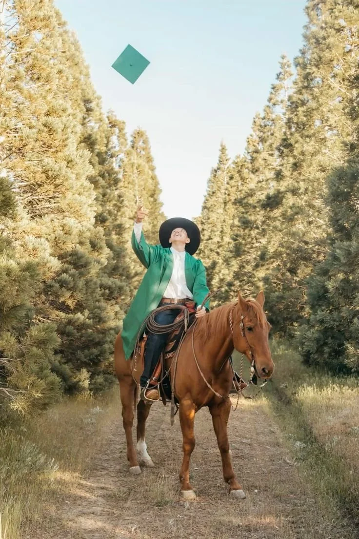 Person wearing a green graduation gown and hat, riding a brown horse through a wooded trail, tossing a hat into the air.