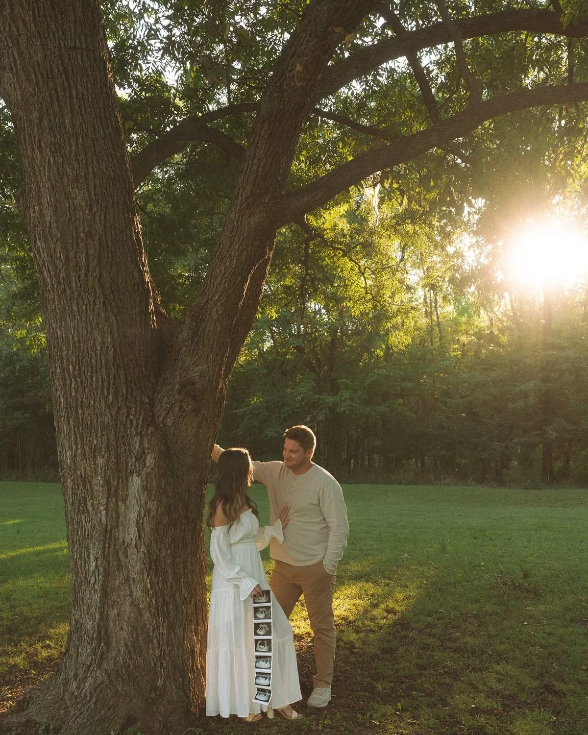 A couple is standing near a large tree in a grassy park, with the sun setting in the background. The woman is wearing a long white dress and holding a small box with ultrasound images inside, while the man is dressed casually in a light shirt and beige pants, gently touching the woman's shoulder and looking at her.