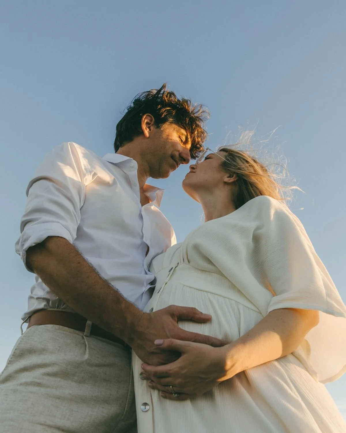 A man and pregnant woman sharing an intimate moment outdoors during sunset, with their foreheads touching.