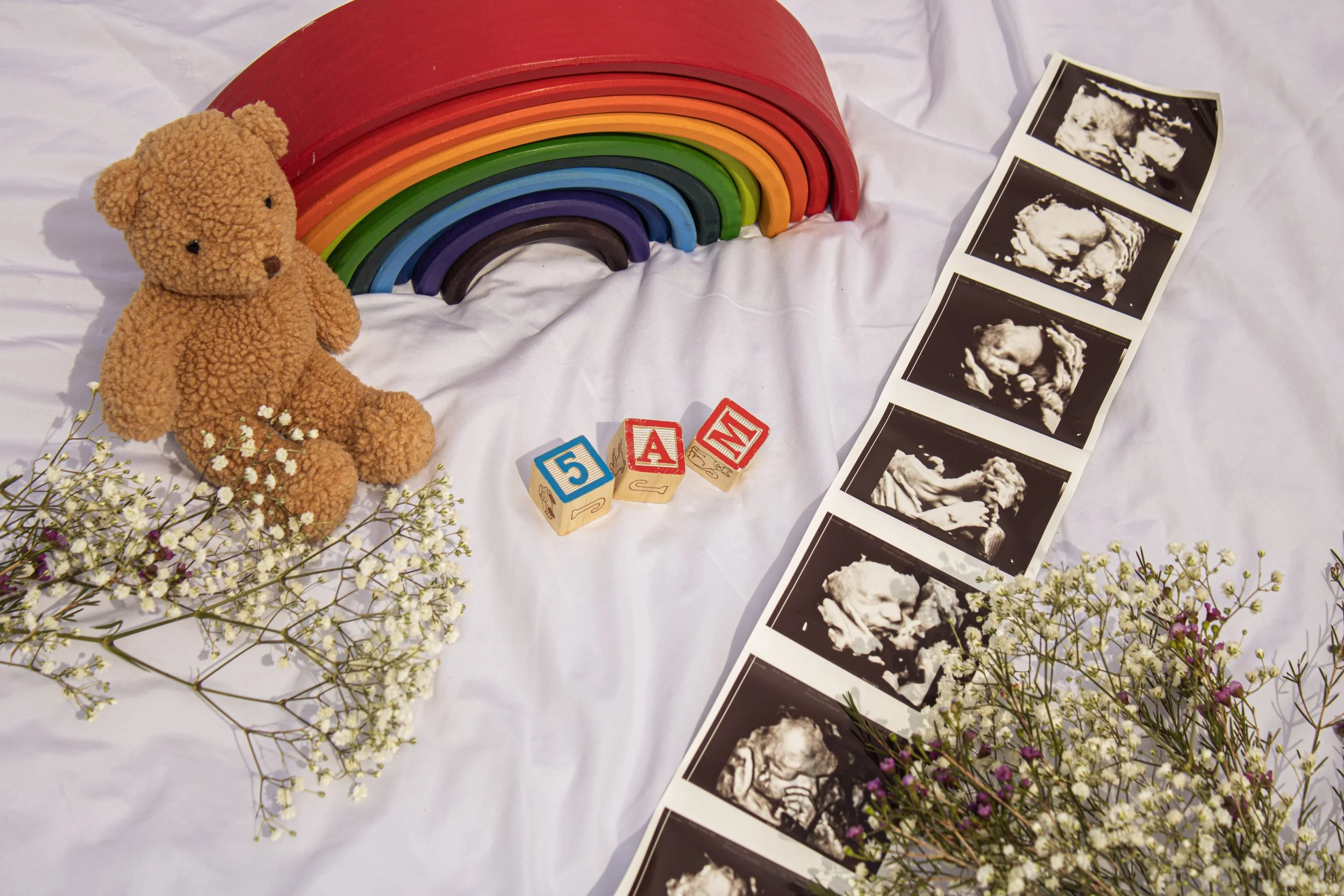 A craft display with a plush teddy bear, a rainbow made of painted wooden arches, baby ultrasound pictures, colorful letter blocks spelling 'M', and flowers arranged on a white fabric surface.