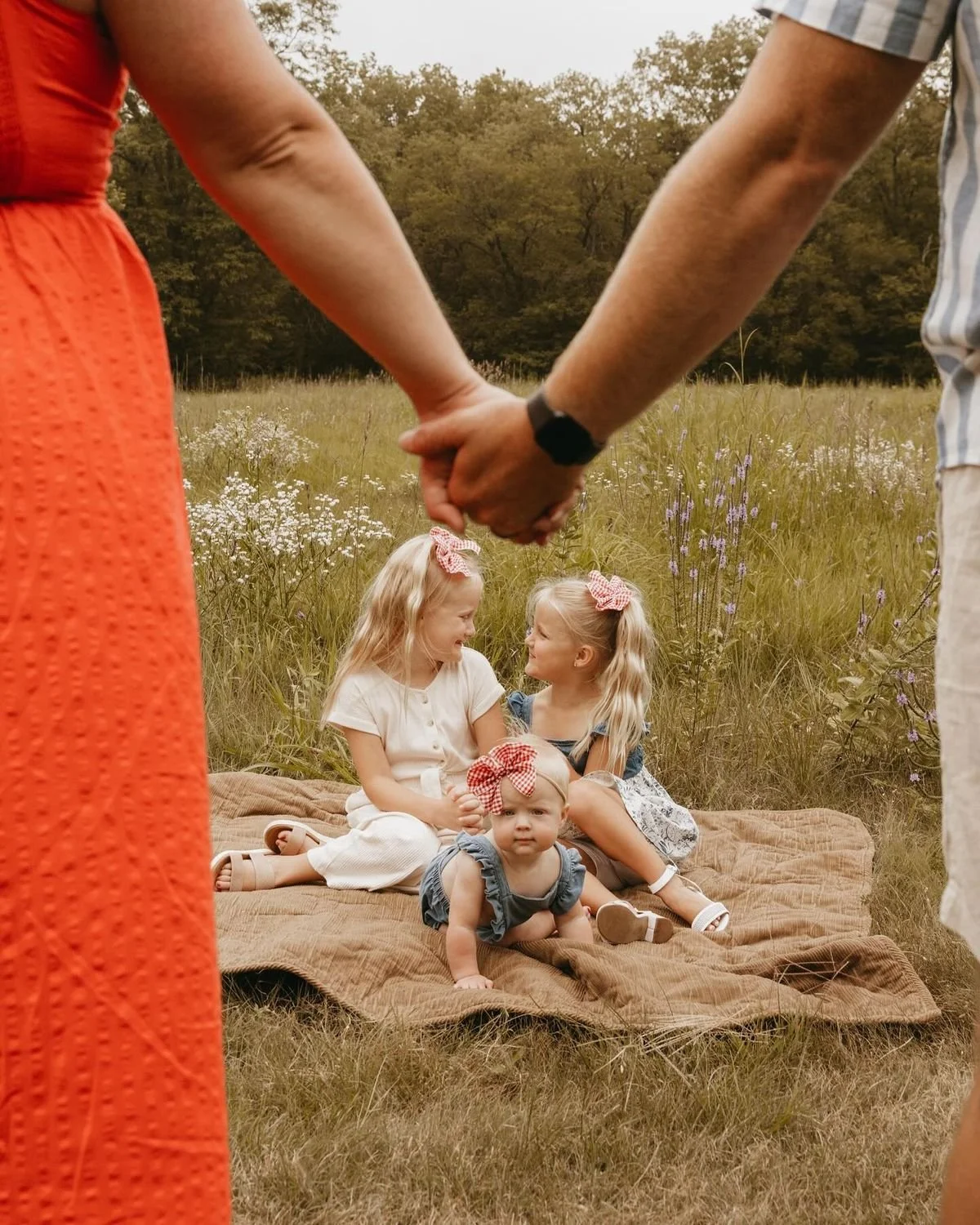 Two adult couples holding hands above three young girls sitting on a blanket in a meadow with wildflowers, autumn trees in the background.