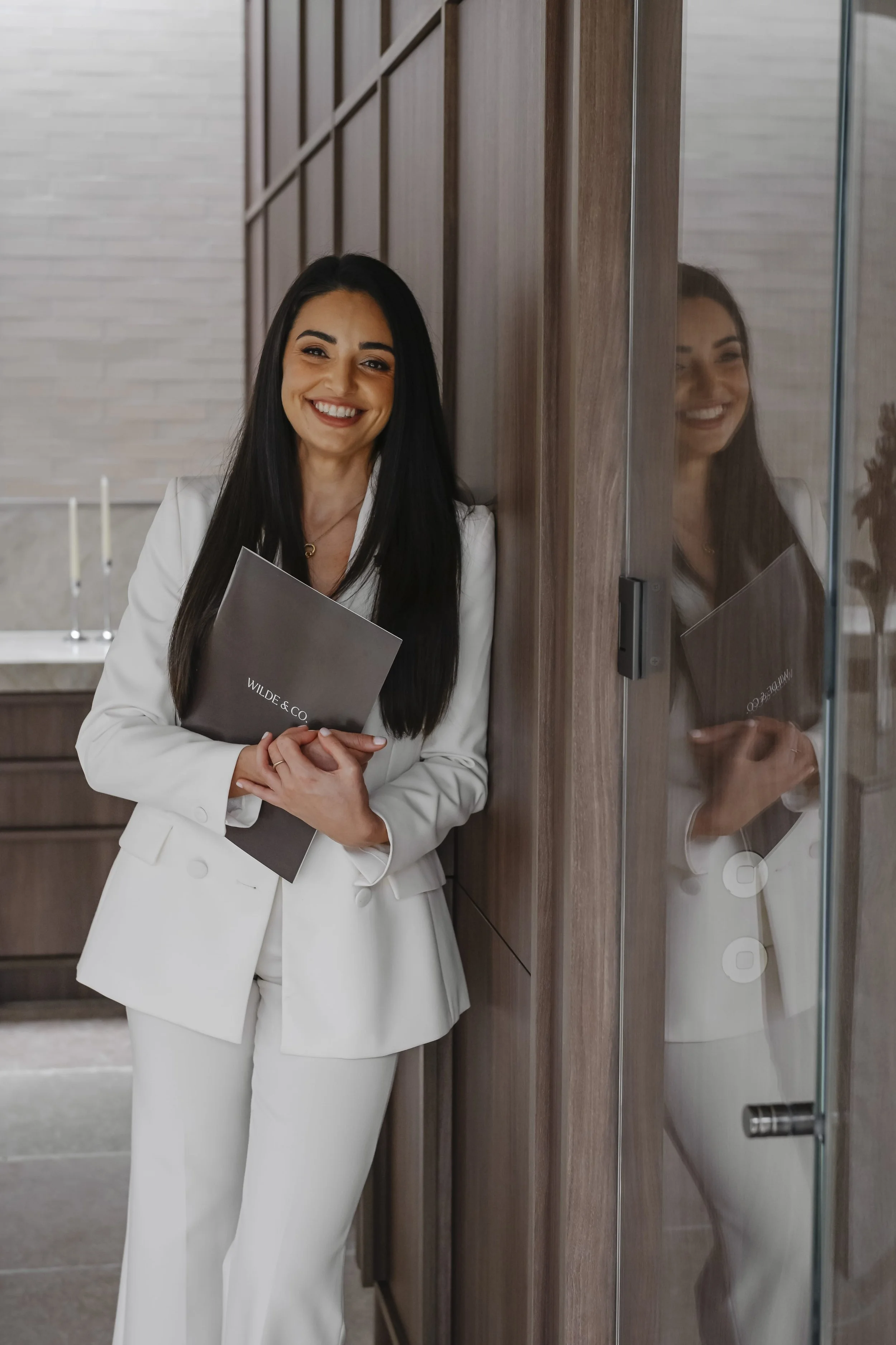 Asli wearing a white suit, smiling and holding a dark gray folder in a modern interior with wood panel walls, standing next to a glass door that shows her reflection.