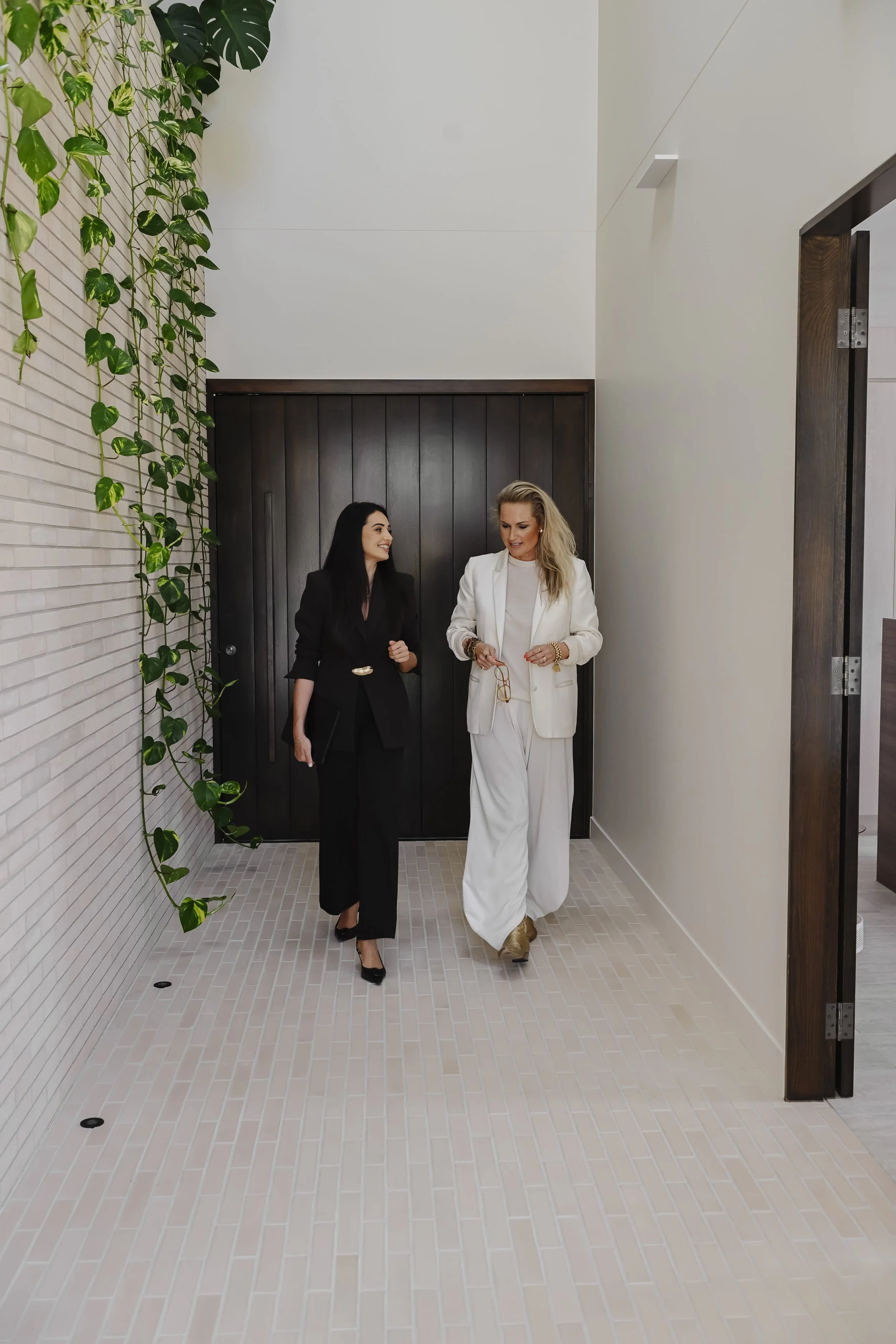 Two women walking and talking in a modern interior hallway with light-colored tile floor, dark wood door, and a vertical garden with green plants on the left wall.