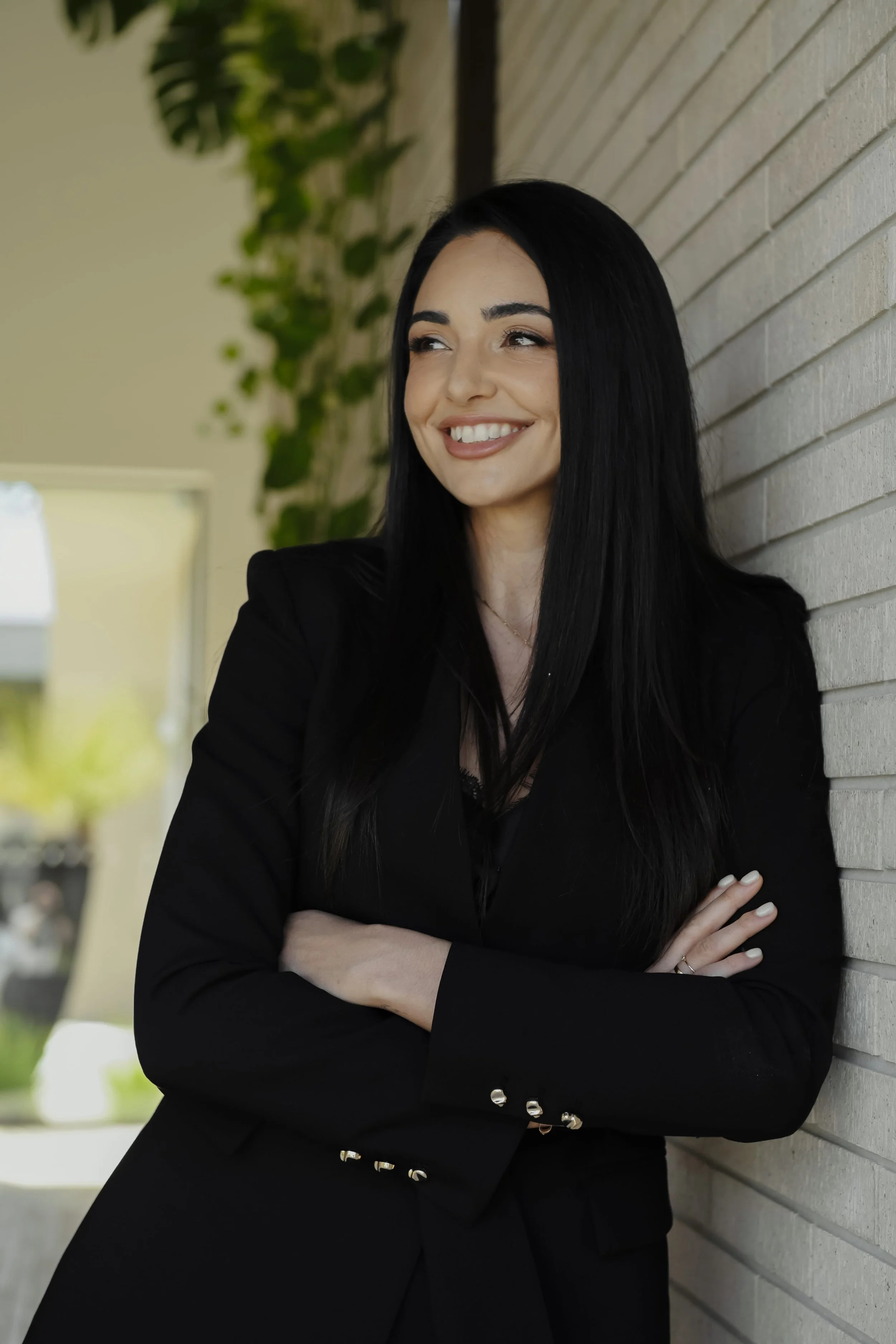 Asli smiling with crossed arms, leaning against a brick wall with a blurred background of green plants and outdoor furniture.