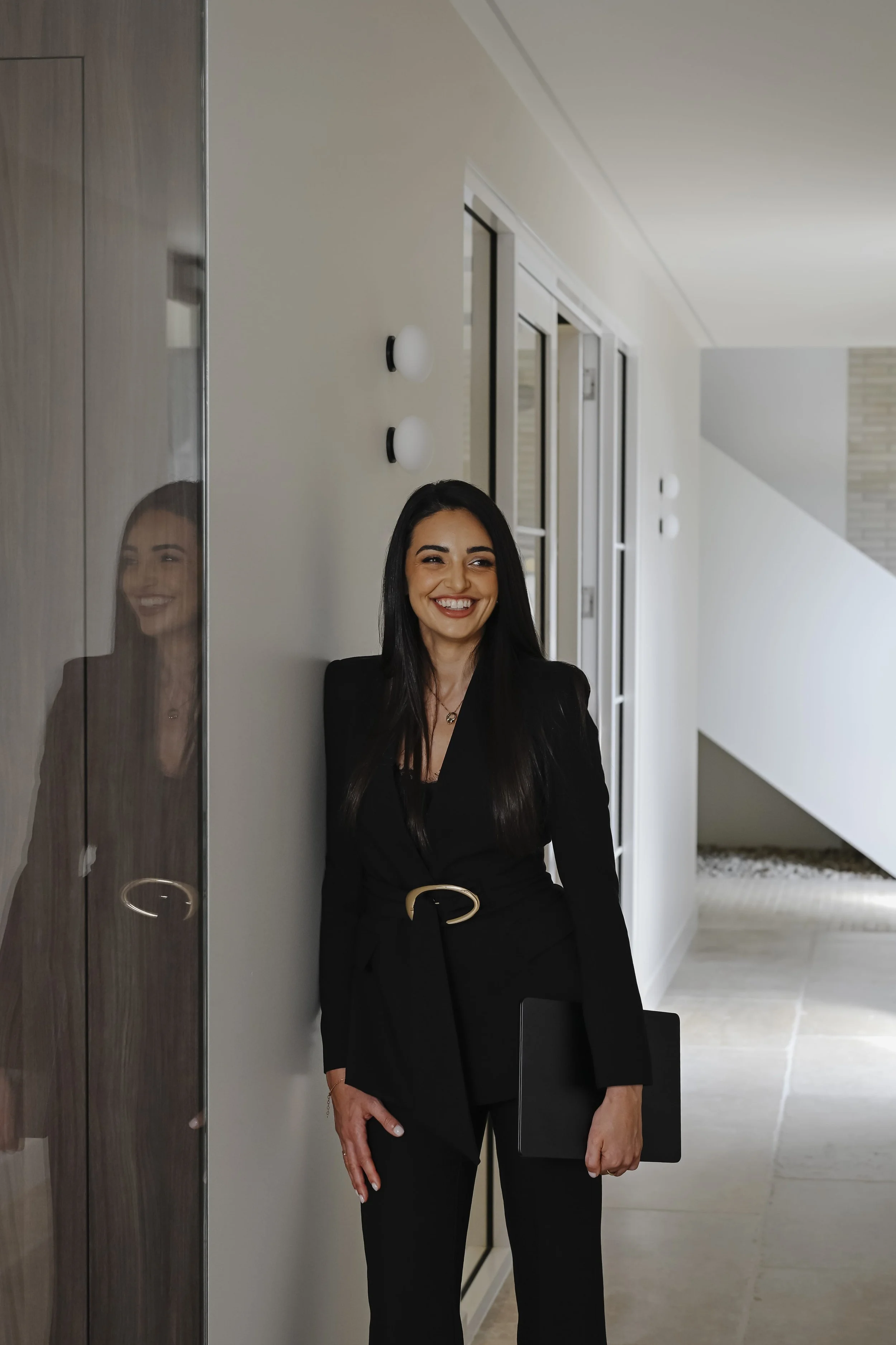 Asli Wilde smiling and standing indoors, with her reflection visible on a glass surface to her left.