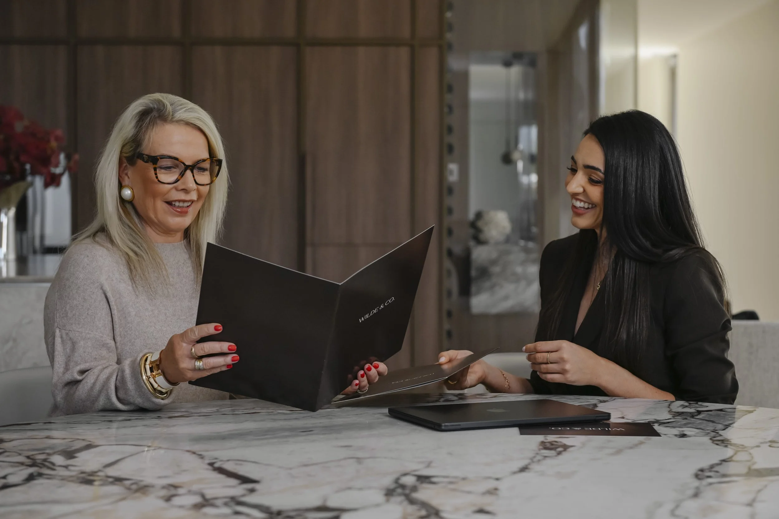 Two women sitting at a marble table in a modern interior, looking at a black folder and smiling, one with blonde hair and glasses, the other with dark hair.
