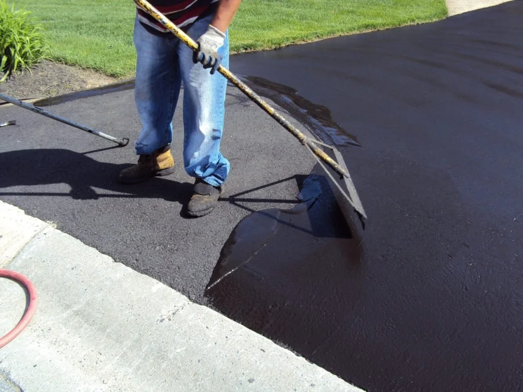 A construction worker is sealing asphalt on a driveway with a long asphalt squeegee.