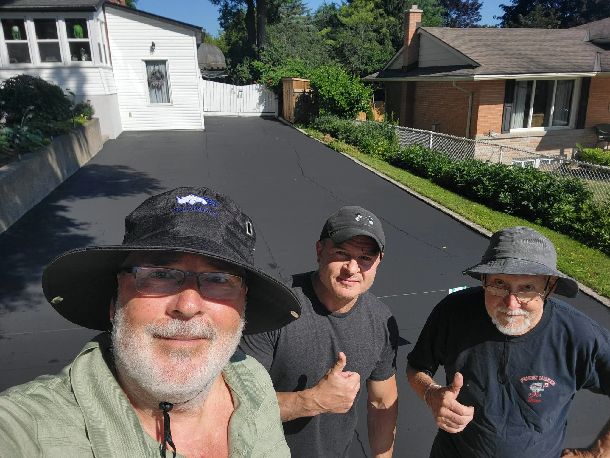 A group of four men with hats and glasses standing outside on a driveway, smiling and giving thumbs-up, with houses and trees in the background.