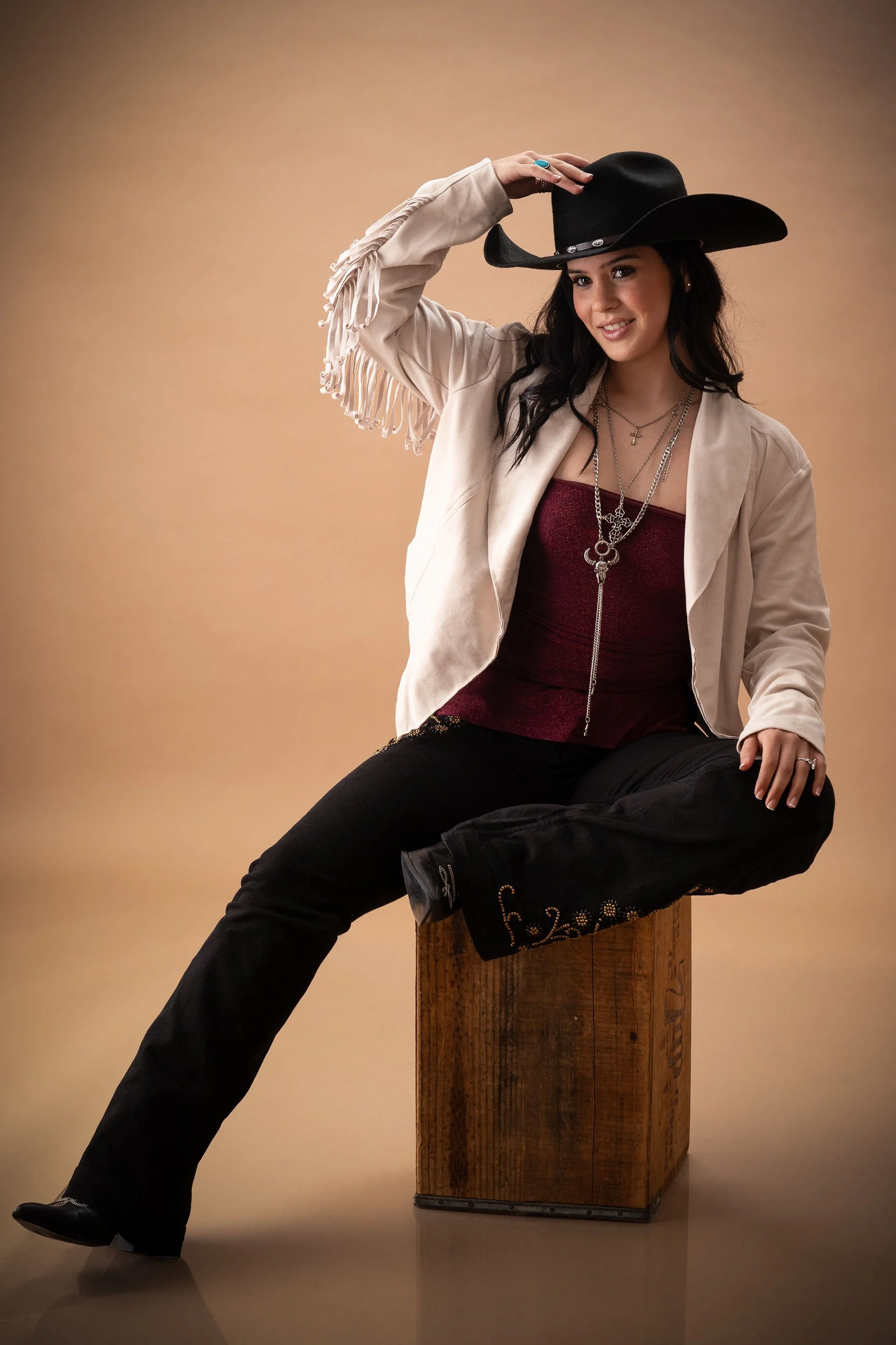 A woman in a beige jacket with fringes, and a red top, sits on a wooden box, wearing a black cowboy hat, black pants with decorative embroidery, and layered necklaces. She is smiling and holding her hat with one hand against a neutral background.